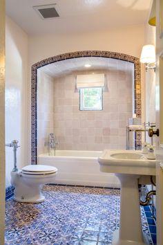 a white toilet sitting next to a bath tub under a window in a bathroom with blue and white tiles on the floor