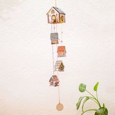 a house shaped mobile hanging from a wall next to a potted plant and white wall