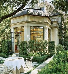 a table and chairs in front of a house with hedges around it, surrounded by trees