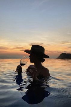 a woman holding a wine glass in her hand while standing in the water at sunset