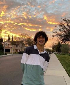 a young man standing on the sidewalk in front of some houses with a sunset behind him