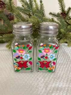 two glass salt and pepper shakers with flowers painted on the sides, sitting next to evergreen branches