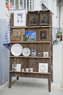 a wooden shelf with pictures and plates on it in an open space between two buildings