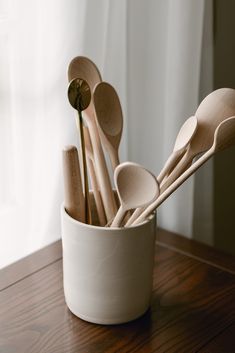 wooden spoons are in a white cup on a wood table next to a window