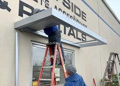 a man standing on a ladder in front of a building while painting the side of a building