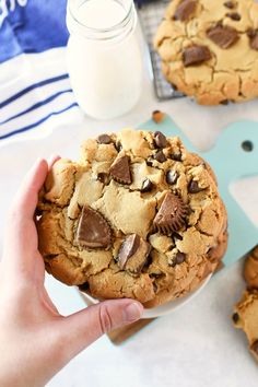 a person holding a chocolate chip cookie in front of other cookies on a table with milk and utensils