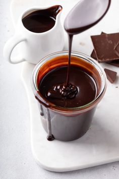 chocolate sauce being poured into a cup with spoons on a white tray next to some pieces of chocolate