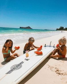 three young children sitting on the beach with a surfboard and watermelon slices