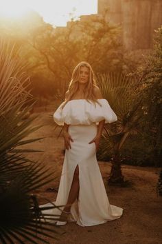 a woman wearing a white dress standing in front of some trees and bushes with her hands on her hips