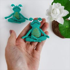 a hand holding two small crocheted frog brooches next to a white flower