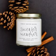 sweet water weather candle surrounded by pine cones and cinnamons on a dark background with the words sweet water weather written in black