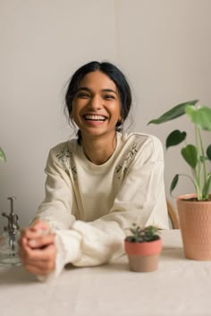 a woman sitting at a table with two potted plants in front of her smiling