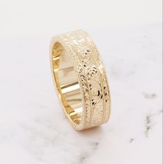 a gold wedding ring with intricate engraving on it's sides, sitting on a marble surface