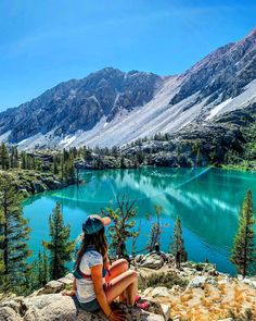 a woman sitting on the edge of a cliff overlooking a lake with mountains in the background