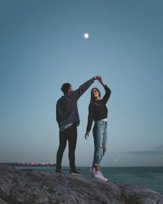 two people standing on top of a rock next to the ocean holding hands and looking at the moon