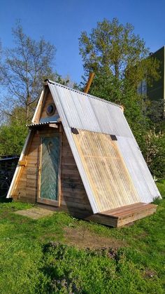 a small wooden structure with a metal roof and door on top of grass in front of trees