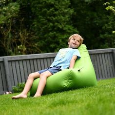 a young boy sitting on a green bean bag chair in the grass with trees behind him