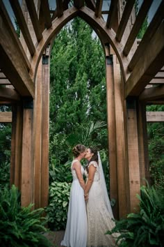 two brides kissing in front of an arch with greenery and trees behind them