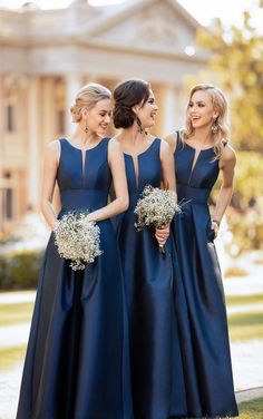 three bridesmaids standing together in front of a large building wearing blue dresses and holding bouquets