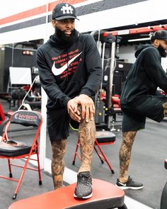 a man with tattoos sitting on top of a red bench in a gym wearing a black hoodie