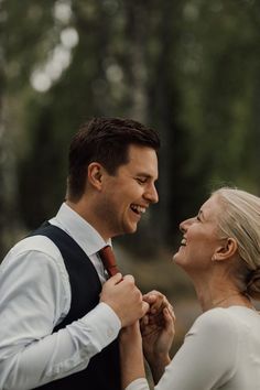 a man and woman standing next to each other in front of trees smiling at each other