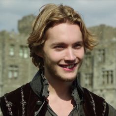 a man with long hair and beard smiles at the camera in front of a castle