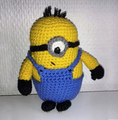 a crocheted yellow and blue minion with big eyes sitting in front of a white wall