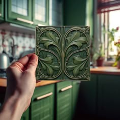 a hand holding up a decorative tile in a kitchen with green cabinets and counter tops