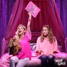 two women sitting on top of a bed holding up a pink book in the air