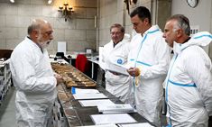 five men in white suits are looking at papers on a long table with donuts