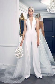 a woman standing in front of a mirror wearing a white jumpsuit and holding a bouquet