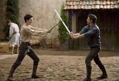 two men are holding swords in the middle of an outdoor area with people standing around