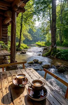 two cups of coffee sit on a wooden table in front of a river and log cabin