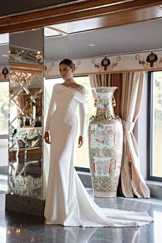 a woman standing in front of a large mirror wearing a white dress and holding a vase