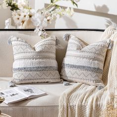 two decorative pillows sit on a couch next to a vase with flowers and a magazine