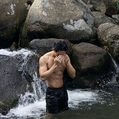 a shirtless man standing in the water next to rocks and holding his hands together