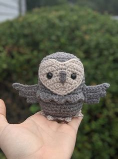 a small crocheted owl sitting on top of a persons hand in front of some bushes