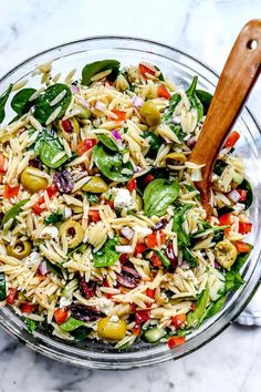 a glass bowl filled with pasta salad on top of a marble countertop next to a wooden spoon