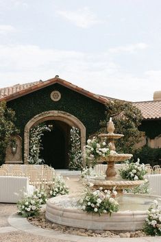 a fountain with flowers on it in front of a building that has an archway and white chairs around it