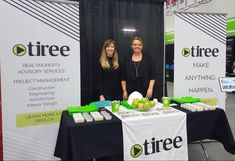 two women standing behind a table with green apples on it at a trade show,