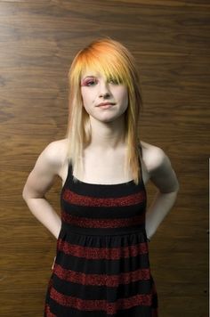 a young woman with yellow hair standing in front of a wooden wall wearing a black and red striped dress