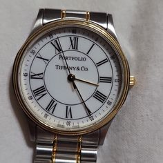 An Attractive 33mm Wristwatch By Tiffany & Co., Portfolio Model. 18k Yellow Gold Tone And Stainless Steel. Roman Numerals. White Dial. Swiss Made Quartz Movement, And The Wristband Has A Deployant Clasp And Fits Up To A 9" Wrist. Men's Size But Great For Women As Well. Was An Award For A Union Pacific Employee For His Services. Very Minor Scratches On The Clasp. Very Clean And Functional. Comes Complete In Display Case With Original Tiffany & Co Box. New Sold At $3000. Tiffany And Co Box, Portfolio Model, White Dial, Steel Watch, Stainless Steel Watch, Roman Numerals, Swiss Made, Display Case, Tiffany & Co.