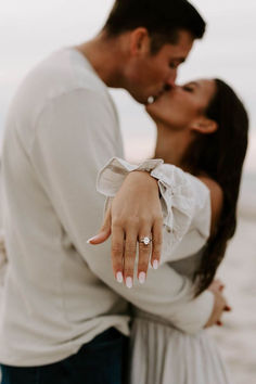 a man and woman kissing on the beach with their engagement rings in their hands as they stand close to each other