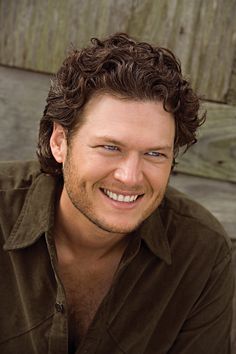 a smiling man with curly hair and brown shirt sitting in front of a wooden wall