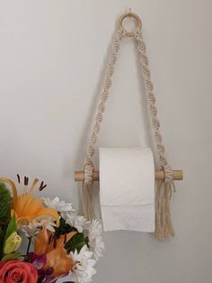 a white toilet paper roll hanging from a rope on a wall next to flowers and a vase with an orange flower in it