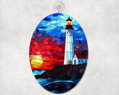 an oval stained glass ornament with a lighthouse on the shore at sunset in front of a red and blue sky