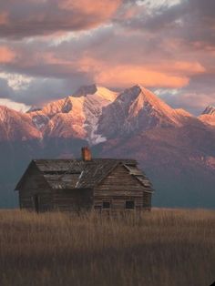 an old house in the middle of a field with mountains in the background at sunset