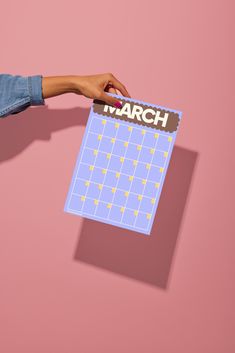 a person's hand holding a march calendar against a pink background