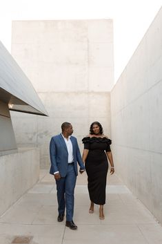 a man and woman are walking down the sidewalk in front of some concrete buildings, one is wearing a black dress