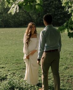 a pregnant woman standing next to a man under a tree in a field with green grass